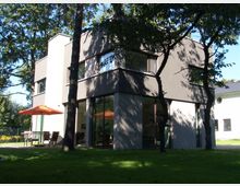 Modernes zweistöckiges Wohnhaus mit großen Fenstern, umgeben von hohen Bäumen und einer Rasenfläche. Im Gartenbereich stehen ein Tisch, Stühle und ein orangefarbener Sonnenschirm.