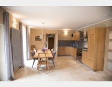 Wohnküche mit moderner Holz-Einbauküche, Eckspüle, Herd und grauer Rückwand. Ein großer Esstisch mit gemischten Stühlen steht im Raum, daneben helle Fliesenböden und bodentiefe Fenster mit grauen Vorhängen.