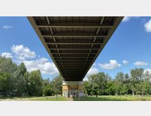 Blick von unten auf eine breite Brücke mit sichtbarer Stahlkonstruktion, die über eine grüne Parklandschaft mit Gras, Bäumen und einem mit Graffiti bedeckten Betonpfeiler verläuft. Der Himmel ist blau mit vereinzelten weißen Wolken.