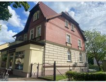 Einfamilienhaus im Stil der Gründerzeit mit roter Klinkerfassade im oberen Bereich und beige verputztem Sockelgeschoss. Das Gebäude hat ein steiles Satteldach mit roten Dachziegeln, zahlreiche Fenster mit dekorativen Verzierungen und eine Metallzaunanlage im Vorgarten.