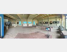 Panoramaaufnahme eines modernen Museumsraums mit Ziegelfußboden und hölzerner Deckenkonstruktion. An den Wänden hängen Informationstafeln und beleuchtete Exponate; in der Mitte befinden sich Vitrinen und Ausstellungselemente.