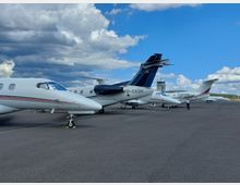 Mehrere Geschäftsflugzeuge stehen nebeneinander auf dem Vorfeld eines Flughafens, mit sichtbaren Fahrwerkskeilen auf dem Asphalt. Im Hintergrund sind ein kleiner Kontrollturm sowie eine flache, grüne Umgebung unter einem wolkigen Himmel zu sehen.