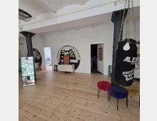 Heller Innenraum eines multifunktionalen Raumes mit Holzboden, weißen Wänden und schwarzen Stützsäulen. An einer Wand befinden sich Pinnwände mit Notizen, daneben ein Boxsack und bunte Hocker; eine offene Tür führt in einen weiteren Raum.