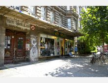 Straßenszene mit einem alten, verzierten Gebäude, dessen Fassade mit Graffiti bedeckt ist, daneben ein Geschäft mit der Aufschrift „Red Star“ und großen Schaufenstern, in denen bunte Kleidung und Dekoration ausgestellt sind. Im Vordergrund stehen parkende Fahrräder und Bäume, die Schatten auf den gepflasterten Gehweg werfen.