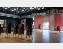 Großer Veranstaltungsraum mit stapelbaren Stühlen auf einem Holzboden, hohen roten Wänden und großen Fenstern auf der rechten Seite, die Tageslicht einlassen. Im Hintergrund ist eine Bühne mit schwarzem Vorhang, darüber Beleuchtung und Technik-Ausrüstung.