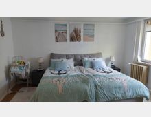 Schlafzimmer mit großem Doppelbett und hellblauer Bettwäsche, flankiert von zwei Nachttischen mit Lampen; darüber hängen drei gerahmte Strandmotive. Rechts befindet sich ein Fenster mit Heizkörper darunter, links steht ein Baby-Hochstuhl mit abgelegten Textilien.