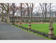 Ein gepflegter Park mit Rasenflächen, gesäumt von Sandsteinstatuen auf Sockeln und großen Laubbäumen, deren Blätter größtenteils gefallen sind. Im Hintergrund steht eine hohe Steinmauer mit Rundbögen, darüber ein Geländer.