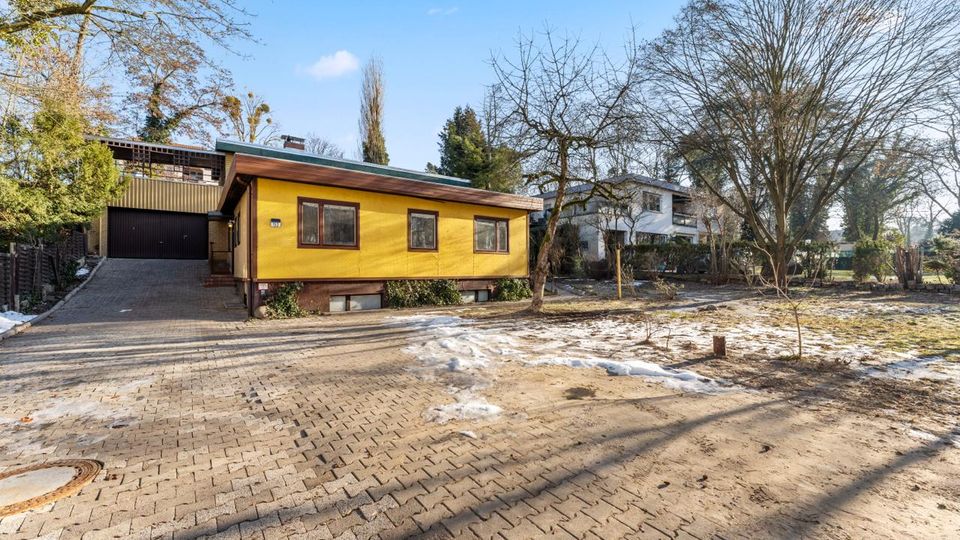 Einfamilienhaus mit gelber Fassade und flachem, weit auskragendem Dach an einer gepflasterten Zufahrt, links mit Garagentor. Davor liegt ein großer, teils sandiger Vorgarten mit kahlen Bäumen und kleinen Schneeresten, umgeben von weiteren Wohnhäusern und Bäumen.