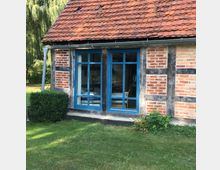 Fachwerkhaus mit einer Fassade aus roten Ziegelsteinen und dunklen Holzbalken, großen blauen Fenstern und einem roten Ziegeldach. Im Vordergrund befindet sich eine gepflegte Rasenfläche mit niedrigem Buschwerk.