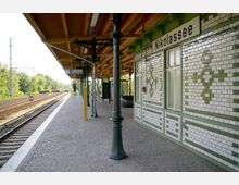 Bahnhofsplattform des S-Bahnhofs Nikolassee in Berlin mit überdachtem Wartebereich, dekorativen gusseisernen Stützen und weißen Fliesen mit grünen Mustern an den Gebäudewänden. Im Hintergrund sind Gleise, ein Signal und umgebende Vegetation sichtbar.