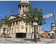 Straßenansicht des Gendarmenmarkts in Berlin mit dem Französischen Dom im Vordergrund, einer klassizistischen Kirche mit Säulen und dekorativen Reliefs. Im Hintergrund ist der Deutsche Dom zu sehen, umgeben von Bäumen, Straßenschildern und Sitzbereichen mit Sonnenschirmen.