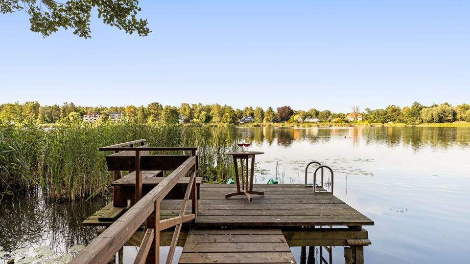 Holzsteg mit Sitzbank und kleinem Tisch führt durch Schilf zu einer Plattform am ruhigen See; seitlich befindet sich eine Metallleiter ins Wasser. Im Hintergrund sind ein bewaldetes Ufer und einzelne Häuser am gegenüberliegenden Seeufer unter klarem Himmel zu sehen.