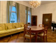 Wohnzimmer mit klassischer Einrichtung: Ein gelbes Sofa vor drei Fenstern mit hellgrünen Vorhängen, ein runder Holztisch mit vier Stühlen, ein Kronleuchter an der Decke, ein Holzschrank in einer Ecke und ein Klavier auf der rechten Seite. Der Boden ist mit einem Parkettmuster gestaltet.