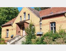 Historisches Gebäude aus gelbem Backstein mit roten Zierbändern und Satteldach, ausgestattet mit rechteckigen Fenstern und einer überdachten Eingangstreppe; der Vorgarten ist teilweise von Büschen und Bäumen bewachsen.