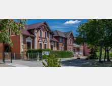 Rotes Backsteingebäude mit weißen Fensterrahmen und Giebeln, umgeben von einer grünen Hecke und einem gepflasterten Weg, mit Bäumen im Vordergrund und blauem Himmel im Hintergrund.