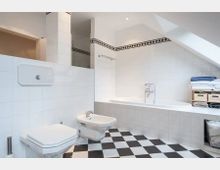 Helles Badezimmer mit weiß gefliesten Wänden, schwarz-weiß kariertem Boden, einer eingebauten Badewanne unter einer Dachschräge, sowie einem WC und einem Bidet. Im Raum befinden sich ein Wandregal mit Handtüchern und Körben sowie ein kleines Fenster, das Tageslicht hereinlässt.