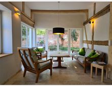 Wohnzimmer mit einer Kombination aus moderner und rustikaler Gestaltung; Holzbalken an den Wänden, große Fensterfront mit Blick auf eine Backsteinmauer und Garten. Eine Couch und zwei Sessel mit grünen Kissen sowie ein Holztisch befinden sich auf einem hellen Steinboden unter einer schwarzen Hängelampe.