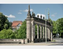 Historische Säulenstruktur mit dekorativen Statuen und Balustraden, umgeben von grünen Bäumen und im Hintergrund ein Haus mit rotem Ziegeldach. Die Umgebung zeigt Bürgersteige, Straßenlaternen und eine angrenzende Straße.