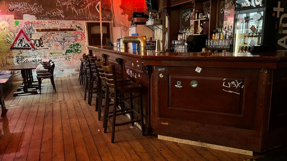 Innenraum einer urigen Bar mit langem dunklem Holztresen, Barhockern und Regalen voller Flaschen und Gläser. Holzdielenboden, beschmierte Wände mit Graffiti sowie ein beleuchteter Getränkekühlschrank und Schilder über dem Tresen prägen den Raum.