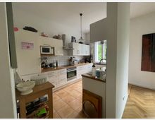 Eine helle, moderne Küche mit weißen Schränken und Holzakzenten. Ausgestattet mit einem Herd, einer Mikrowelle, einem Kühlschrank und einer Spüle unter einem Fenster; der Boden ist mit beigen Fliesen ausgelegt, angrenzend ist ein Bereich mit Holzboden sichtbar.