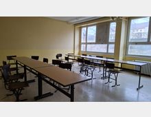 Leerer Klassen- oder Seminarrraum mit mehreren langen Tischen und einzelnen Stühlen, beige gestrichenen Wänden und linoleumbedecktem Boden. Große Fenster entlang einer Wand lassen Tageslicht herein; darunter stehen Heizkörper und sichtbare Rohrleitungen verlaufen an Decke und Wand.