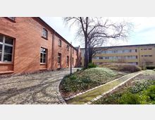Backsteinbau mit mehrteiliger Fassade und großen Fenstern auf der linken Seite, daneben eine gepflasterte Gehwegfläche und eine begrünte Gartenanlage mit einem großen, kahlen Baum in der Mitte. Im Hintergrund ist ein modernes, beige-gelbes Gebäude mit schmalen Fenstern zu sehen.