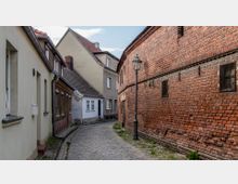 Schmale gepflasterte Gasse mit einer Mischung aus alten Backsteinhäusern und verputzten Gebäuden, die teils sichtbare Abnutzung zeigen. Ein historischer Laternenpfahl steht inmitten der Straße, und die Ziegelwand rechts dominiert die Szenerie.