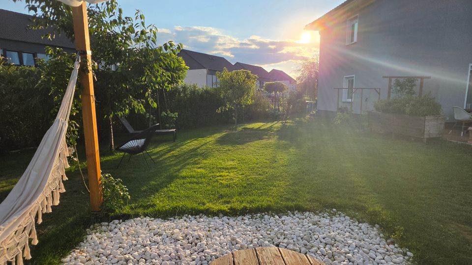 Garten eines Wohnhauses mit großer Rasenfläche, kleinen Bäumen und Hecken, im Vordergrund ein Kiesbett und ein Holzpfosten mit aufgehängter Hängematte. Rechts steht das Haus mit Terrasse und Hochbeet, im Hintergrund weitere Wohnhäuser im Abendlicht.