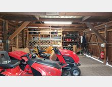 Innenansicht einer Holzgarage mit offenem Dachgebälk, in der rote Aufsitzmäher im Vordergrund stehen. Im Hintergrund befinden sich offene Regale mit Werkzeugen, Materialien und Kanistern sowie ein Fahrrad, eine Feuerlöscherstation und weitere gelagerte Gegenstände.