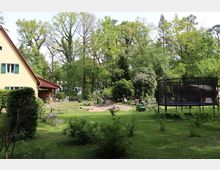 Großer Garten mit einer Rasenfläche, vereinzelten Büschen und Bäumen im Hintergrund. Im linken Bereich steht ein gelbes Haus mit grünen Fensterläden und einem Holzunterstand, rechts ist ein Trampolin und eine kleine Holzhütte zu sehen.