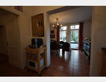 Wohnraum mit Essbereich und Küche, gekennzeichnet durch Terrakottafliesen, einen Holzrollwagen mit Kaffeemaschine und Mikrowelle sowie einen Essbereich mit dunklen Holzmöbeln vor großen Fenster- und Türflächen, die den Blick auf einen winterlichen Garten freigeben. Ein Kronleuchter hängt von der Decke, und rote Vorhänge rahmen die Fenster.