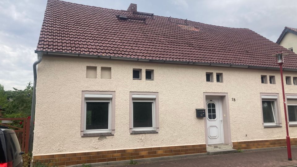 Einfamilienhaus mit heller Putzfassade, roten Dachziegeln und mehreren rechteckigen Fenstern. Der Eingang hat eine weiße Tür und daneben einen Briefkasten; der Bürgersteig davor ist mit Pflastersteinen ausgelegt.