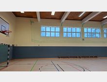 Innenansicht einer Sporthalle mit Spielfeldmarkierungen, Basketballkorb und Handballtor an der Wand. Hohe Fensterreihen mit Schutznetz, Deckenbalken mit Leuchten sowie eine lange Holzbank entlang der blaugrau gestrichenen Wand.