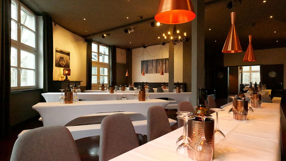 Innenraum eines modernen Restaurants mit langen, weiß gedeckten Tischen, gepolsterten Stühlen und großen Fenstern entlang der linken Wand. Über den Tischen hängen kupferfarbene Pendelleuchten; im Hintergrund sind Säulen, Wandkunst und weitere Tischreihen zu sehen.