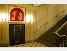 Inneneinrichtung eines historischen Gebäudes mit einer grünen Treppe rechts, verziertem Geländer und hellbeigen Wänden mit goldfarbenen Streifen. Über einem dekorativen Lüftungsgitter ist eine rote Wandnische mit antikisierenden Figuren zu sehen, flankiert von zwei Wandlampen mit warmem Licht.