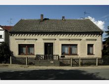 Einstöckiges Wohnhaus mit Putzfassade, rechteckigen Holzfenstern und Satteldach, umgeben von einem niedrigen Lattenzaun, mit Eingangstür in der Mitte und einem kleinen Garten davor. Der Himmel ist klar und blau im Hintergrund.