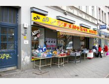 Fassade eines kleinen Discounters mit gelbem Schild „Schnäppchen Markt“ und dem Hinweis „ab 49 Cent“. Vor dem Geschäft stehen mehrere Metallkörbe mit Waren, darunter Spielzeug und Haushaltsartikel.