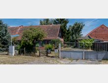 Backsteinbauernhaus mit Ziegeldach und grünen Fensterläden, umgeben von Bäumen und Sträuchern. Vor dem Haus befindet sich ein Metalltor und ein kleiner Holzzaun, mit einem Straßenschild "Alter Wittenberger Weg" im Vordergrund.