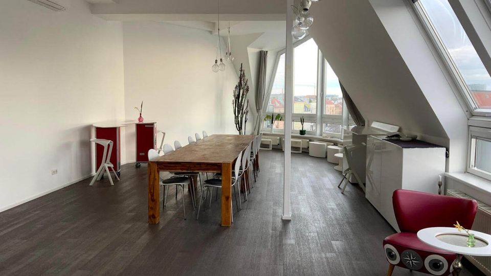 Bright loft-style meeting room with a long wooden table and chairs, white walls, and hanging pendant lights. Slanted ceiling with large skylight windows and a seating area by tall windows overlooking nearby rooftops.