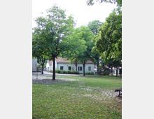 Grüne Parklandschaft mit vereinzelten Bäumen und einer gepflasterten Fläche, im Hintergrund ein Gebäude mit hellen Wänden, roten Ziegeldächern und mehreren Fenstern. Rechts im Vordergrund steht eine einfache Parkbank.