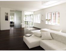 Modernes Wohnzimmer mit weißem Sofa und dunklem Holzboden, großen Fenstern, die viel Tageslicht hereinlassen, und einer Glastür, die zu einem angrenzenden Raum führt. Auf einem Fensterbrett steht eine Vase mit Blumen, die Gesamtgestaltung wirkt offen und minimalistisch.