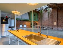 Industriell gestalteter Wohnraum mit offener Küche und Essbereich, sichtbaren Backsteinwänden, Holzdecke und einer grünen Zwischengalerie mit Treppe. Hängende Pflanzen und breite Fenster sorgen für ein loftartiges Ambiente.