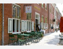 Straßenszene mit rotem Backsteingebäude, das ein Café und weitere Geschäfte beherbergt. Vor dem Café stehen grüne Tische und Stühle auf einem gepflasterten Gehweg, daneben ein Eingang mit Speisekarte und Blumenarrangement.