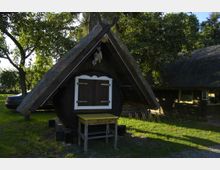 Ein kleines Holzgebäude in Form eines Fasses mit einem steilen, mit Reet gedeckten Dach, einem weißen Fensterladen und einem gelben Holztisch davor, umgeben von Bäumen und grünem Rasen. Im Hintergrund sind weitere Gebäude mit Reetdächern sichtbar.