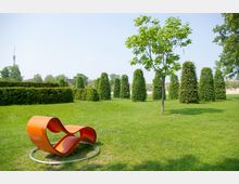 Grüne Parklandschaft mit einer modernen, orangefarbenen Skulptur in geschwungener Form auf einer Rasenfläche. Im Hintergrund sind geschnittene Hecken und kegelförmig getrimmte Büsche zu sehen, ergänzt durch vereinzelte Bäume.