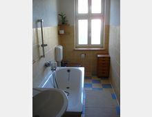 Schmales Badezimmer mit gelb-beigefarbenen Fliesenwänden und blau-gelben Bodenfliesen. Ausgestattet mit einer Badewanne, einem Waschbecken, einem kleinen Holzschrank, einem Fenster mit mattiertem Glas und einem Handtuchhalter.