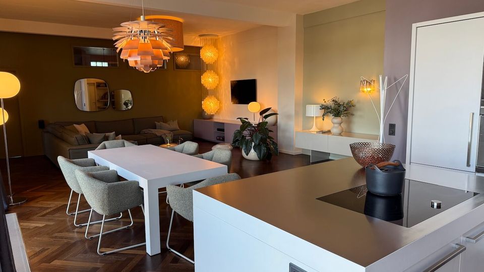 Open-plan modern kitchen and dining area with a white island countertop and built-in cooktop, leading to a dining table with upholstered chairs and a lounge with a sectional sofa and wall-mounted TV. The space features dark herringbone wood flooring, pendant and globe lighting, muted green and beige walls, and decorative mirrors and plants.