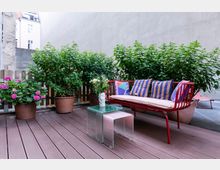 Kleine Terrasse mit Holzdielenboden, einem roten Sofa mit bunten Kissen, einem gläsernen Beistelltisch und Pflanzen in Terrakotta-Töpfen, umgeben von hohen Hauswänden. Im Hintergrund sind teilweise Fassaden angrenzender Gebäude sichtbar.