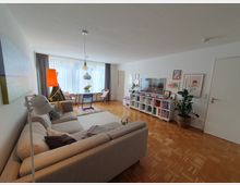 Wohnzimmer mit Holzboden und hellen Wänden, eingerichtet mit einem großen beigen Sofa, einem Esstisch mit Stühlen vor einer großen Fensterfront mit hellen Vorhängen, einem Bücherregal mit dekorativen Elementen, einem Fernseher und Zimmerpflanzen. Wandbilder und Lampen ergänzen die gemütliche Einrichtung.