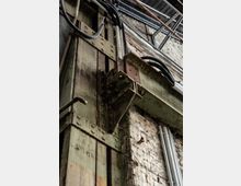 Detailaufnahme einer verwitterten alten Industrieanlage, die eine rostige Metallkonstruktion mit Schrauben und Nieten zeigt. Im Hintergrund sind eine ausgeblichene Ziegelwand und Kabelleitungen zu sehen.